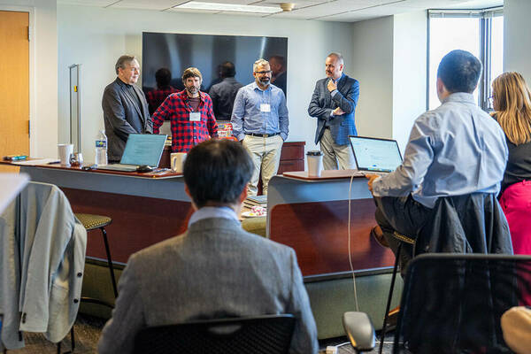 Four men stand in front of a meeting held in a casual setting.