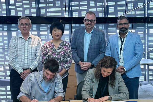 Two individuals sign documents at separate tables while four people stand behind them observing. The backdrop is a modern architectural wall featuring a geometric pattern.  The scene appears to be a formal agreement signing.
