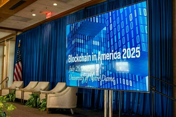 A large screen displays the title "Blockchain in America 2025," with the date, July 25, 2025, and location, University of Notre Dame, beneath it.  The background of the screen shows an abstract representation of binary code in shades of blue.  In front of the screen are several empty beige armchairs and potted plants, with a US flag to the left and blue drapes in the background.