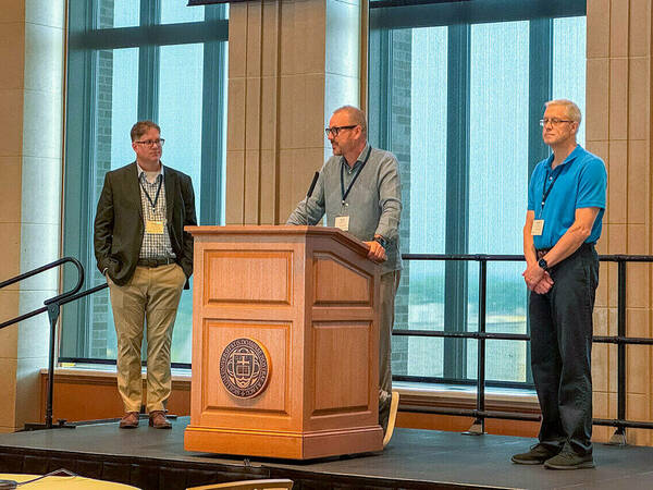 Three men stand on stage, one behind a podium, speaking to an audience.