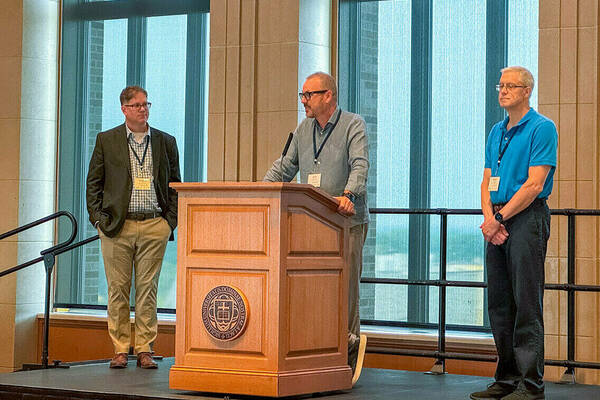 Three men stand on stage, one behind a podium, speaking to an audience.